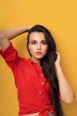 Cheerful mixed race brunette in red blouse isolated on yellow background. Beauty surprised woman with long dark hair and clean skin looking at camera while touching her hair with hands