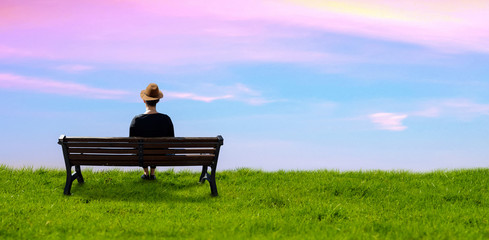 a hat wear girl sitting on a bench in a meadow green grass background sky.