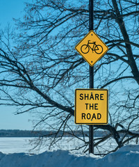 SHARE THE ROAD bicycle sign posted in a park, with tree branches, blue sky and a snow covered lake in the background, on a sunny, picturesque winter day.