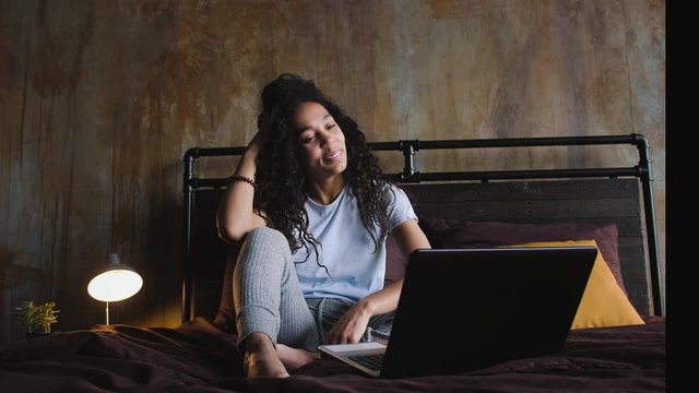 Young attractive African-American woman using laptop computer for a video call in the bed, waving hands, slow motion. Work from home concept