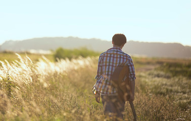 Asian boy holding guitar, walking on the country hill with dry grass. Back view of dreamer musician