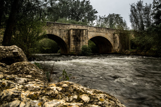 Puente desde la orilla del r&iacute;o un d&iacute;a nublado en larga exposici&oacute;n.