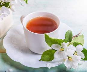 Green tea in white cup with twig of blossoming apple tree on shaped saucer on wooden table. Apple holiday. Apple day. Tea time. Closeup.
