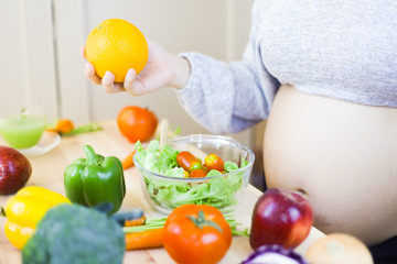 Pregnant women wear long sleeved gray sweaters. She is holding mandarin oranges and putting a salad on a glass on the cooking table. The table is full of fresh vegetables. Mummy nutrition for pregnant