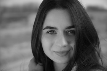 Black and white bright portrait of young white skin Caucasian woman looking directly to camera, holding her hair, which wind blowing around, and smiling. Beautiful youth. Blurry bokeh background.