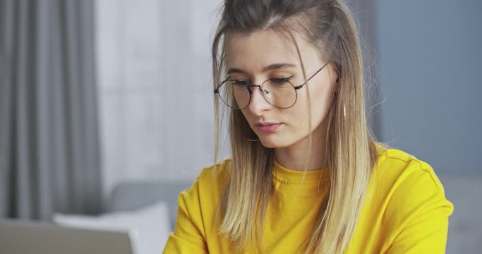 Confident young woman working at a computer, checking email and responding online. The girl finishing work and adjusting glasses, smiling happily and looking at the camera.