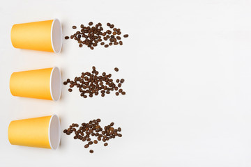 Three yellow paper cups and splashes of coffee beans on a white background. Flatlay. Coffee to go concept. Copy space on the right.