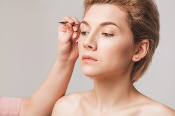 Naklejka premium the browist makes correction with black tweezers. Beautiful blonde model close-up on a beige background in the studio. Skin retouching