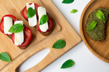 Traditional Italian Caprese bruschetta: sandwich toast with basil pesto sauce, mozzarella cheese and tomatoes on the wooden board surrounded by basil leaves and tomatoes