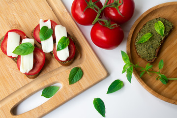 Traditional Italian Caprese bruschetta: sandwich toast with basil pesto sauce, mozzarella cheese and tomatoes on the wooden board surrounded by basil leaves and tomatoes