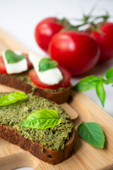 Traditional Italian Caprese bruschetta: sandwich toast with basil pesto sauce, mozzarella cheese and tomatoes on the wooden board surrounded by basil leaves and tomatoes