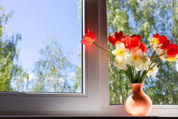 Bright red white and yellow flowers in a vase stand on a white windowsill. Outside the window is green foliage of trees. sunlight falls through the window