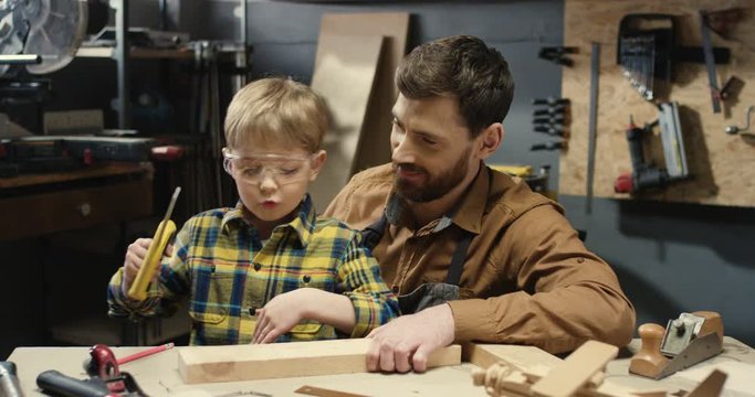 Little Caucasian cute boy in goggles sawing timber in workshop at his father carpenter. Dad teaching his small son to file wood and giving five to each other. Successful work concept.