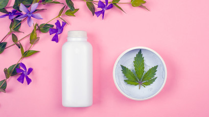 white bottles with cosmetics on a pink background with a leaf of marijuana and violet wildflowers top view. beauty, skin care