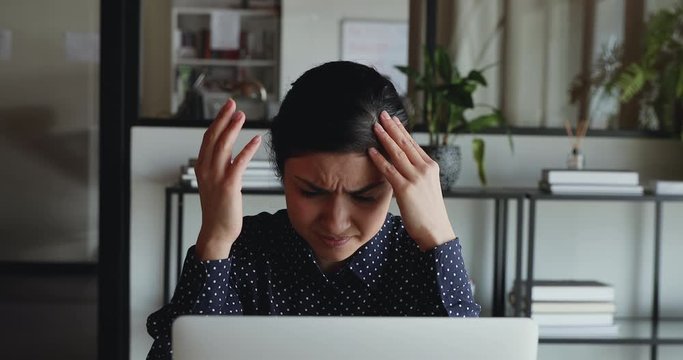 Frustrated overwhelmed young indian business woman feels headache or stress working on laptop computer sitting in office. Upset overworked female worker looking desperate about migraine at workplace.