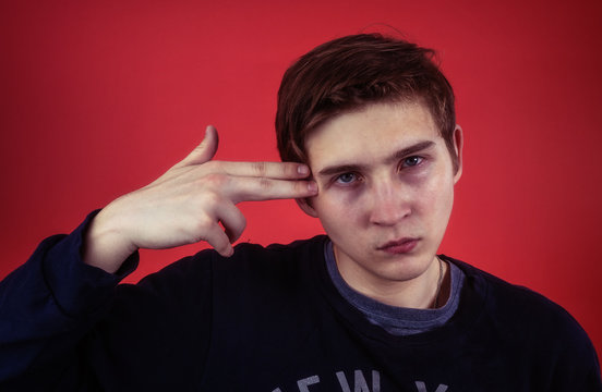 Depressed young male keeping hand gun near temple and looking at camera while pretending to commit suicide against red background