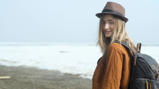 travel along the seacoast. Portrait of young Stylish beautiful hipster girl in casual clothes in a hat with a backpack on her back against the background of sandy empty beach. Active weekend in nature