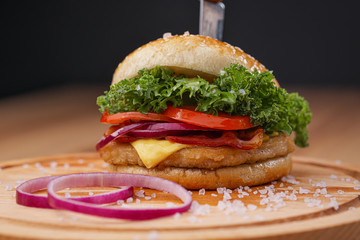 Delicious juicy hamburger on a served wooden board with a stuck knife, on a black background. Beef patties, vegetable buns and chopped onion.