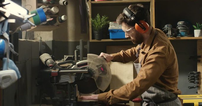 Young Caucasian hansome male carpenter in goggles and headphones cutting wooden plank with a circular electric saw in carpentry workshop. Man joiner proceeding wood.