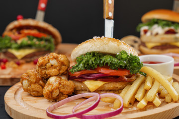 Combo set of fast food. Juicy hamburger, French fries, chicken legs and sauce. Served wooden board and cutlery on a black background. Fast food restaurant. Food delivery.