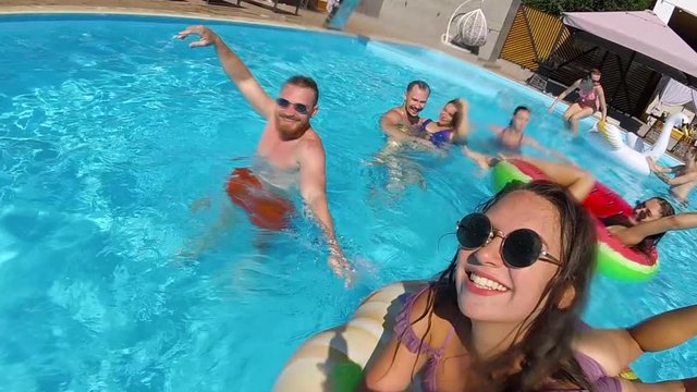 Lifestyle blogger woman taking selfie video with action camera floating on mattress in swimming pool. Travel vlogger girl films vlog at resort. Female live streaming beach party with friends. Slomo.