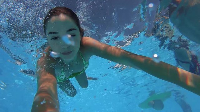 Fitted pretty woman in swimsuit diving under water in swimming pool, shooting herself on action camera. Mixed race girl filming selfie video bathing underwater. Partying with friends on beach party.