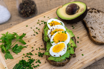 A slice of bread with avocado and eggs on a wooden board. Also salt, chives, parsley, half of avocado and another slice of bread. There is a sackcloth in the background