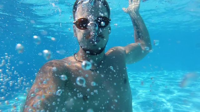 Lifestyle blogger man taking selfie video with action camera diving under water and having fun in swimming pool. Travel vlogger films vlog from party with friends at luxury resort. Slow motion.