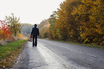Lonely guitarist looking at empty country road in autumn.