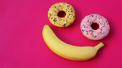 Two donuts and a banana lie on the pink surface, forming an abstraction of the emoticon - smile. Taken from the top point