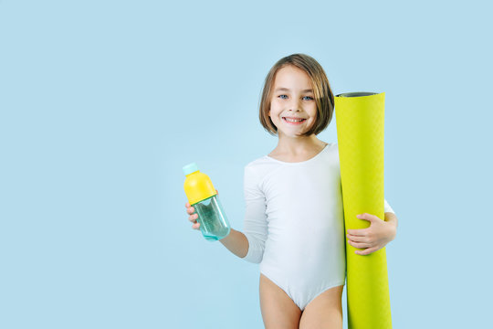 Happy smiling girl in a white leotard with rolled mat and water bottle over blue