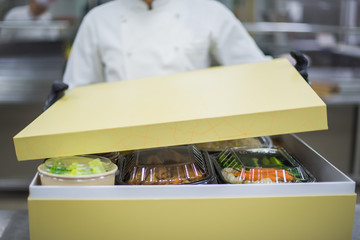 
A chef in black gloves and a white uniform is packing food delivery for customers in a large brown cardboard box.