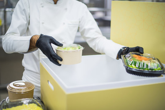 
A chef in black gloves and a white uniform is packing food delivery for customers in a large brown cardboard box.