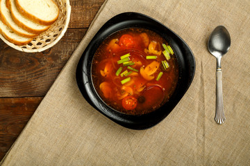 black dish of italian tomato soup with potatoes and mushrooms on a wooden table on a napkin and a spoon and bread.
