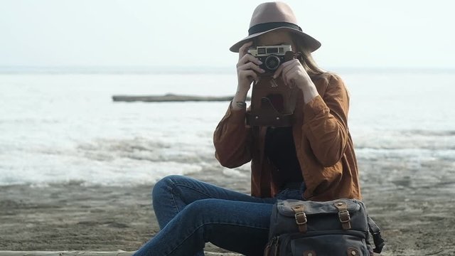 young happy hipster girl in shirt and jeans and stylish hat is sitting on sand by sea with retro camera. girl takes pictures on old camera and laughs. Creative walk along the sea coast for photography
