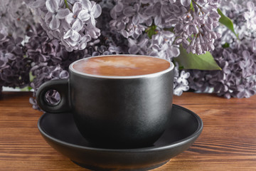 Cup of hot coffee with milk on wooden table, beautiful background with lilacs flowers