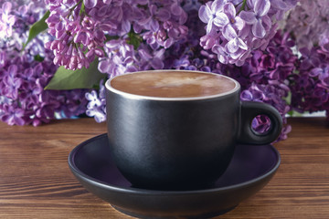 Cup of hot coffee with milk on wooden table, beautiful background with lilacs flowers