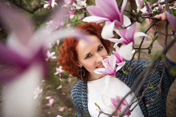 Fototapeta premium Portrait of young pretty red haired woman near blooming magnolia tree on sunny spring day
