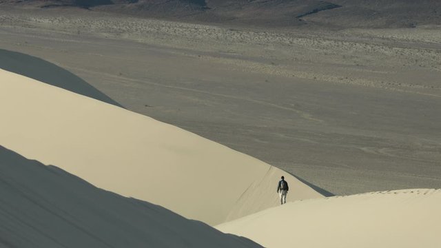 A male hiker walks across the ridgeline of the Eureka Dunes at sunrise in Death Valley National Park in California. The Eureka Valley and Saline Mountain range is seen in the background.