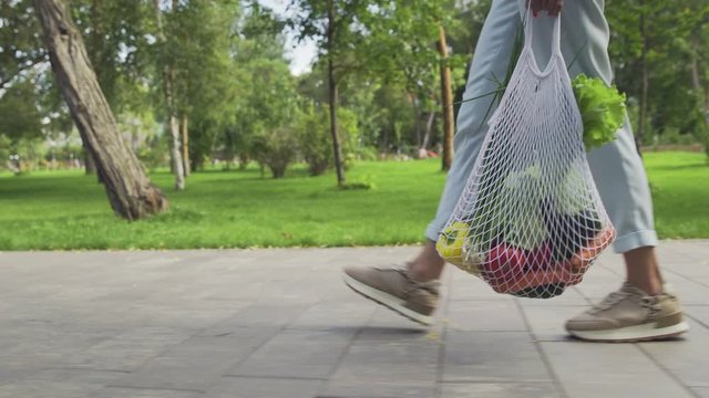 Unrecognizable woman holds cotton mesh shopping bag with vegetables and walks in the summer park