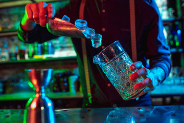Close up of professional barman finishes preparation of alcoholic cocktail in multicolored neon light, gives it to client. Entertainment, drinks, service concept. Modern bar, trendy neoned colors.