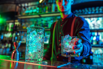 Close up of professional barman finishes preparation of alcoholic cocktail in multicolored neon light, gives it to client. Entertainment, drinks, service concept. Modern bar, trendy neoned colors.