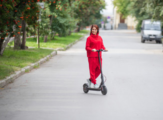 Red-haired girl in a red tracksuit drives an electric scooter. A young woman in oversized clothes rides around the city on modern transport and listens to music using wireless headphones.