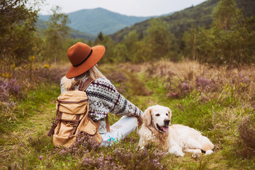 Young hippie blond hair woman in wool sweater with textile backpack and smiling happy golden retriever dog sitting on grass in summer mountains valley. Pets hiking and weekend activities concept. - Powered by Adobe