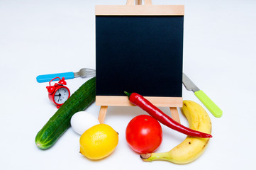 Slate, tomatoes, banana, red paper, lemon, egg, fork, clock, knife and cucumber on white background. Cooking at home. Cooking. Vegetables and fruits. Place for text. Ingredients. Shopping. Copy space.