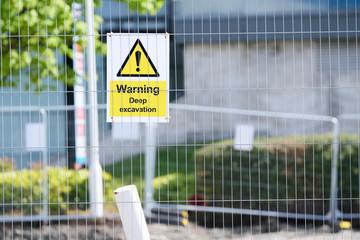 Deep excavations danger sign on fence with trench hole in background on construction site