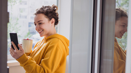 Smiling young woman making video call with smartphone near window balcony terrace at home. Caucasian millennial girl in yellow hoodie holding cup of coffee making selfie, sharing data on social 