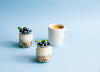 Homemade natural yogurt with granola and blueberries in glass jars and coffee on blue background. Healthy breakfast set.