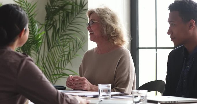 Head shot happy beautiful middle aged businesswoman feeling excited about educational lecture in office. Smiling pleasant senior elder female employee having fun, talking to diverse young colleagues.