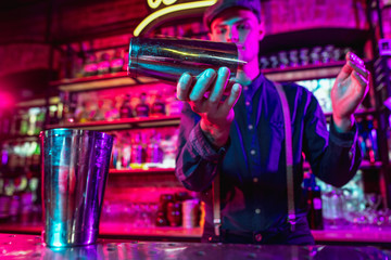 Professional barman finishes preparation of alcoholic cocktail for guest in multicolored neon light. Entertainment, drinks, service concept. Modern bar, crafted beverages, trendy neoned colors.